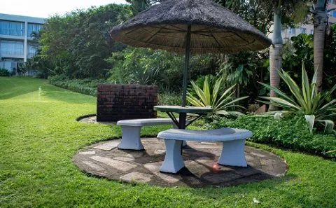 Outdoor seating area with a thatched umbrella surrounded by greenery and a building