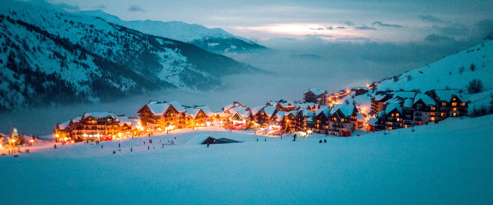 Snowy village with illuminated buildings at dusk surrounded by mountains