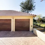 Two wooden garage doors on a house with a brick driveway and garden