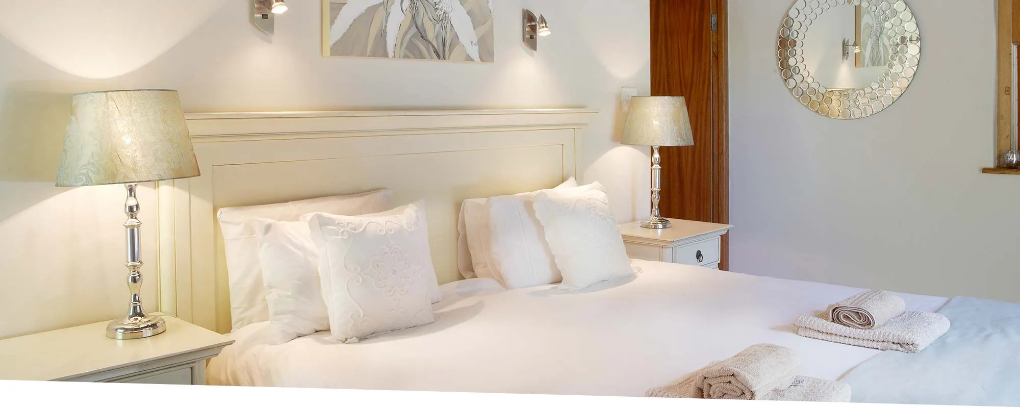 White bedroom with large bed two lamps and a round mirror on the wall