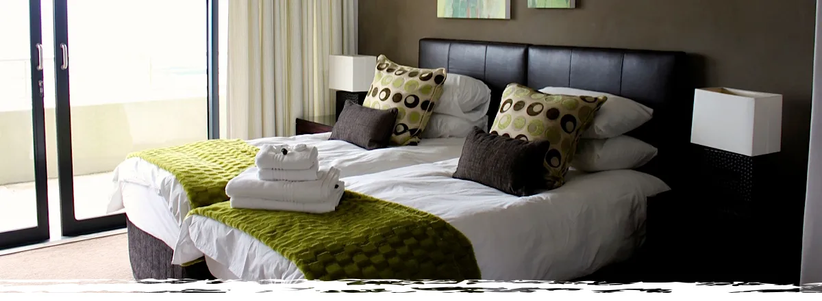 Two beds with green and brown pillows in a modern bedroom with large windows