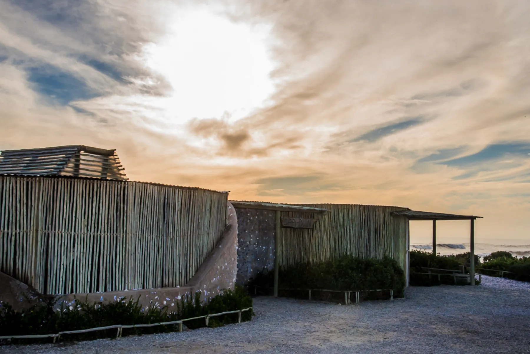 Beach huts with thatched roofs and greenery under a cloudy sky at sunset