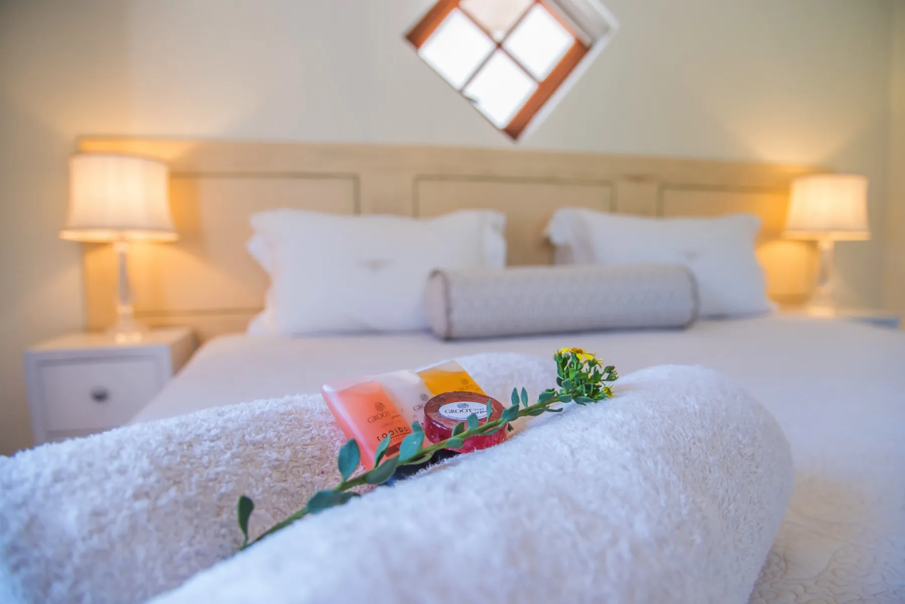 White bed with rolled towel toiletries and flowers on it two bedside lamps