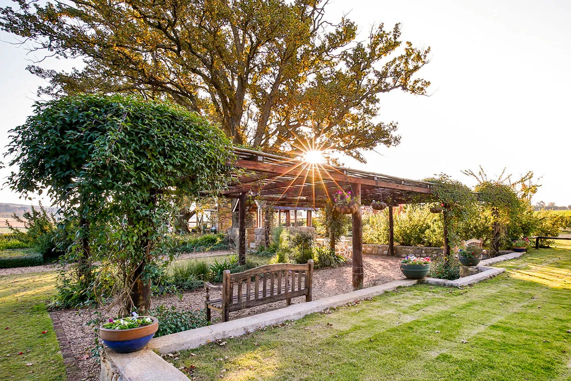 Wooden pergola with bench in a lush garden sun setting in the background
