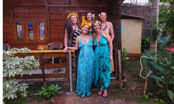 Five women in colorful dresses posing in front of a wooden cabin with plants