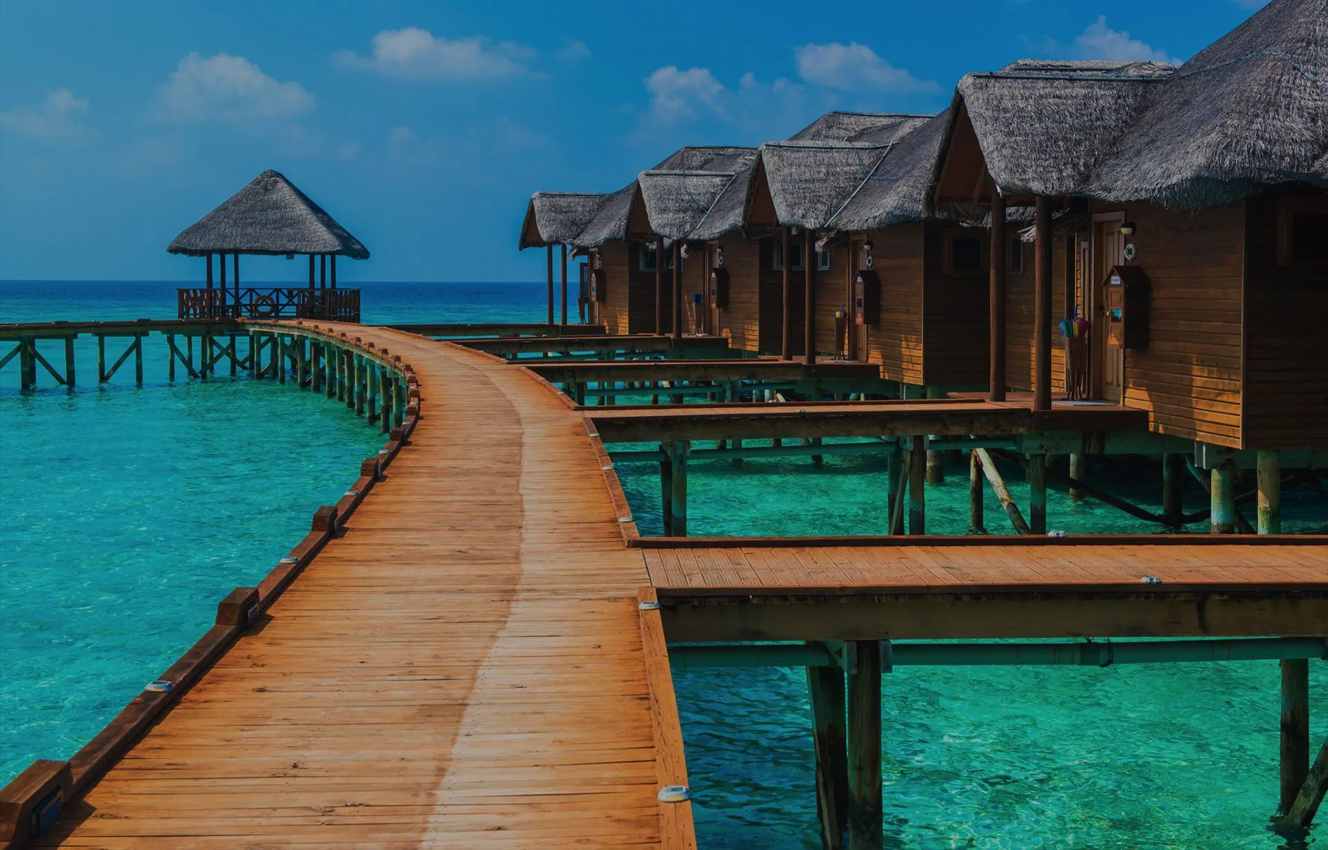 Wooden walkway over water leading to thatchedroof bungalows on stilts in turquoise sea