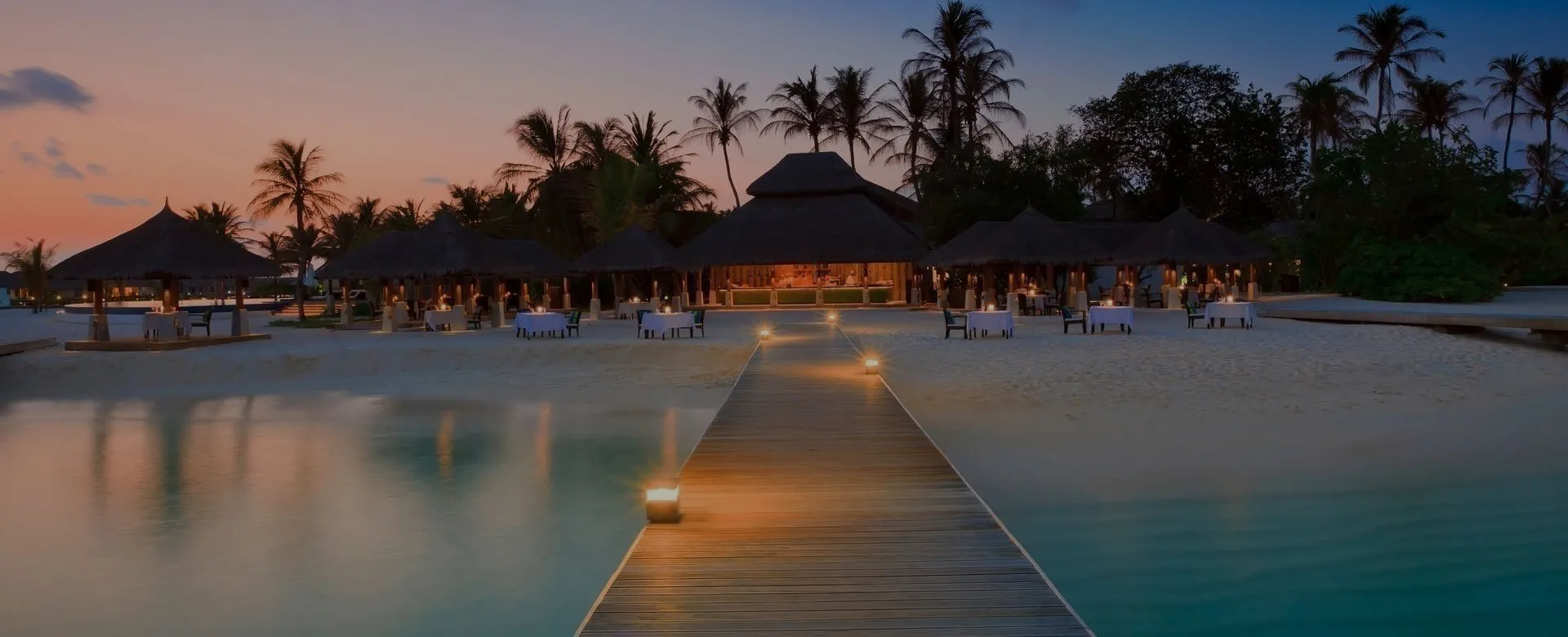 Tropical resort at sunset with a wooden walkway leading to thatchedroof buildings