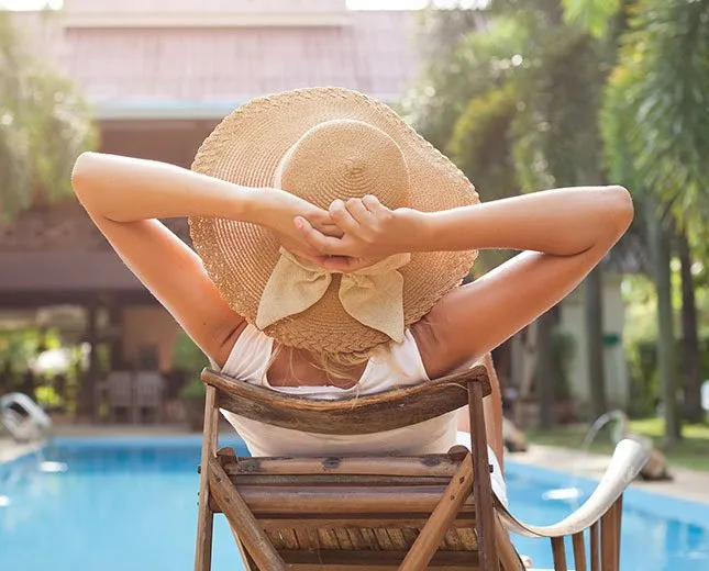 Woman in hat relaxing on chair by pool with arms behind head