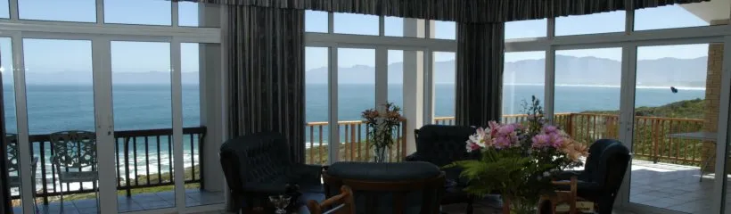 Sunlit room with large windows overlooking the ocean furnished with chairs and flowers