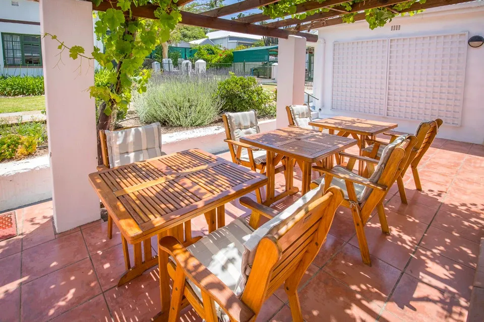 Wooden chairs and tables on a patio with greenery and a white wall