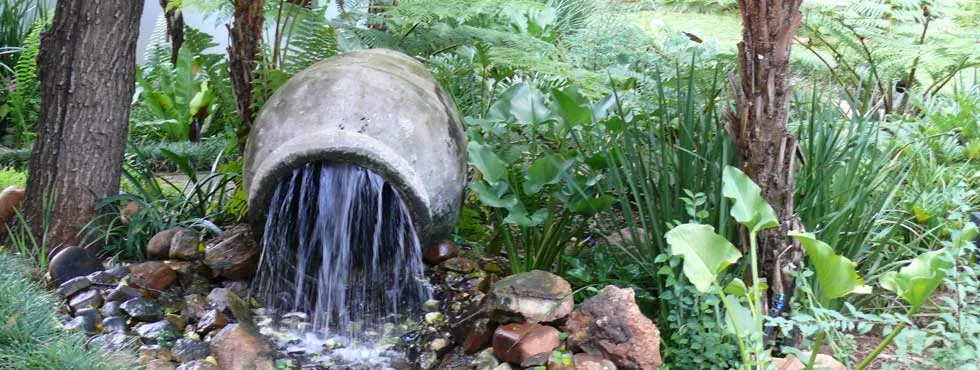 Water flowing from a large pot into a rocky stream surrounded by lush greenery