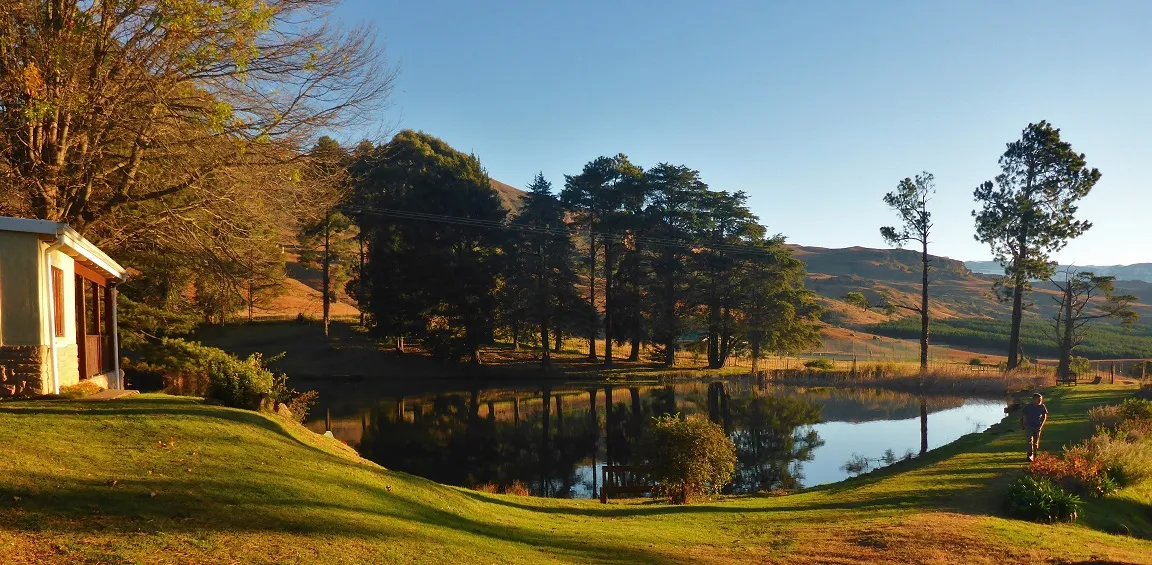 Small cabin by a pond with trees and hills in the background at sunset