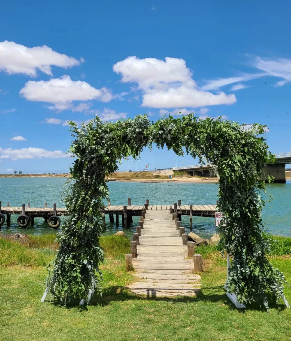 Arch of greenery over wooden path by water with bridge in background