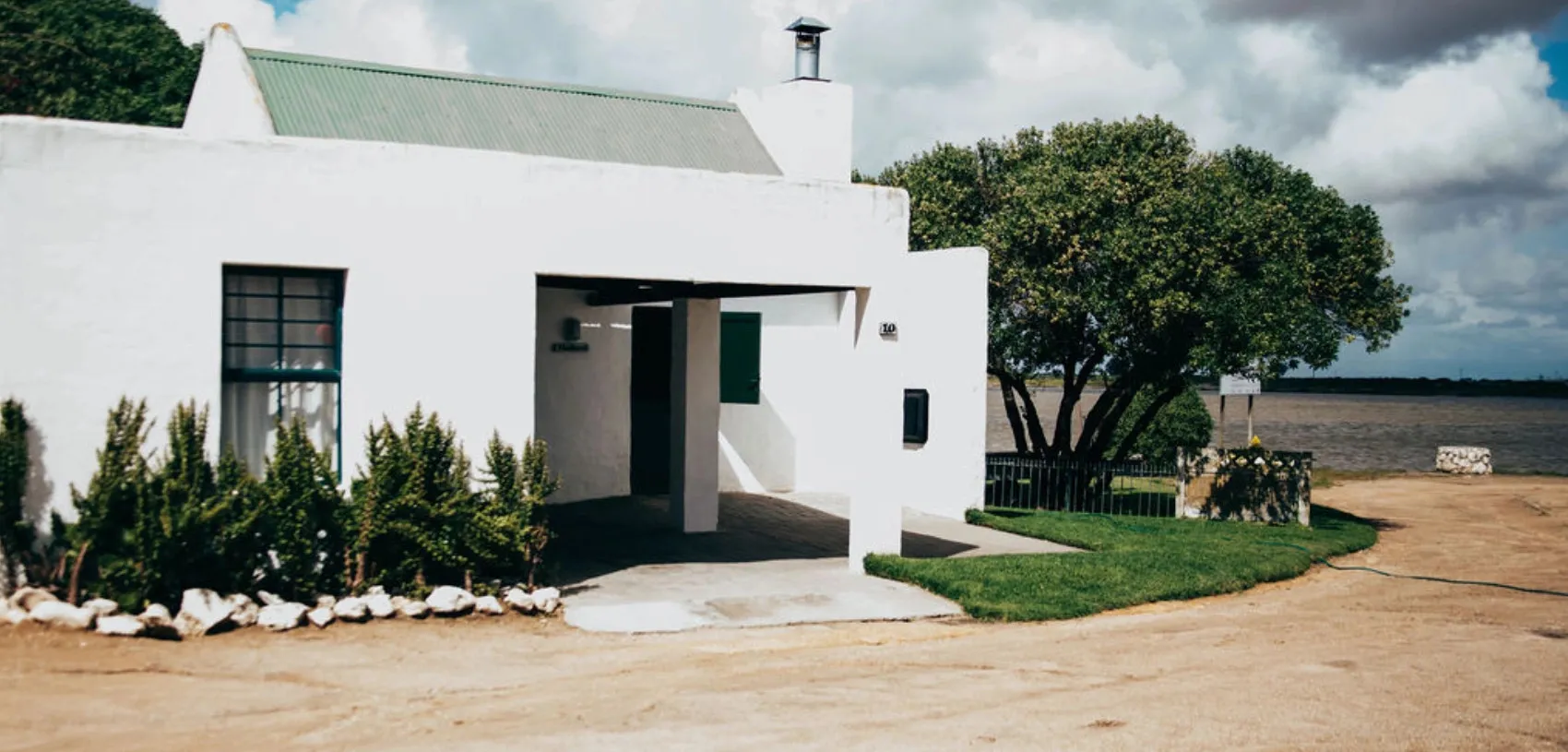 White house with green roof near water surrounded by greenery and a dirt path