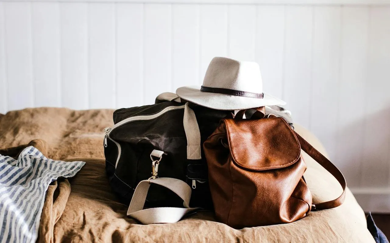 Brown leather backpack and black duffel bag on a bed with a hat on top