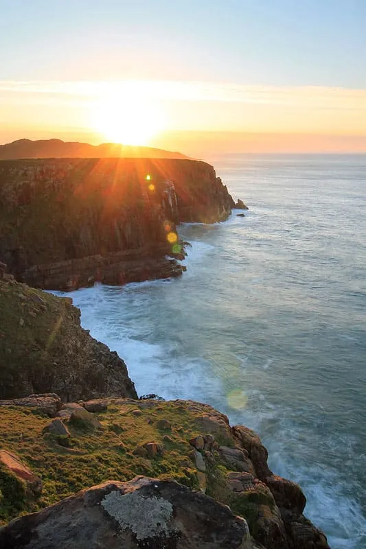 Sunset over ocean with rocky cliffs and waves crashing against them