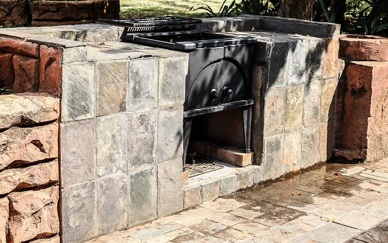 Outdoor stone barbecue grill with black metal grates and lid surrounded by tiles