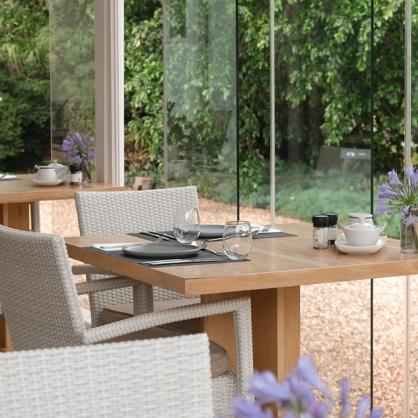 Wicker chairs and set table in a sunlit glassenclosed patio with greenery outside