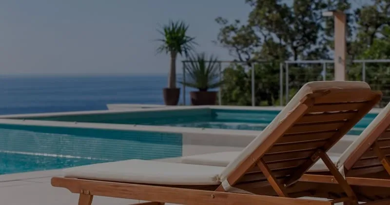 Two wooden lounge chairs by a pool overlooking the ocean with palm trees
