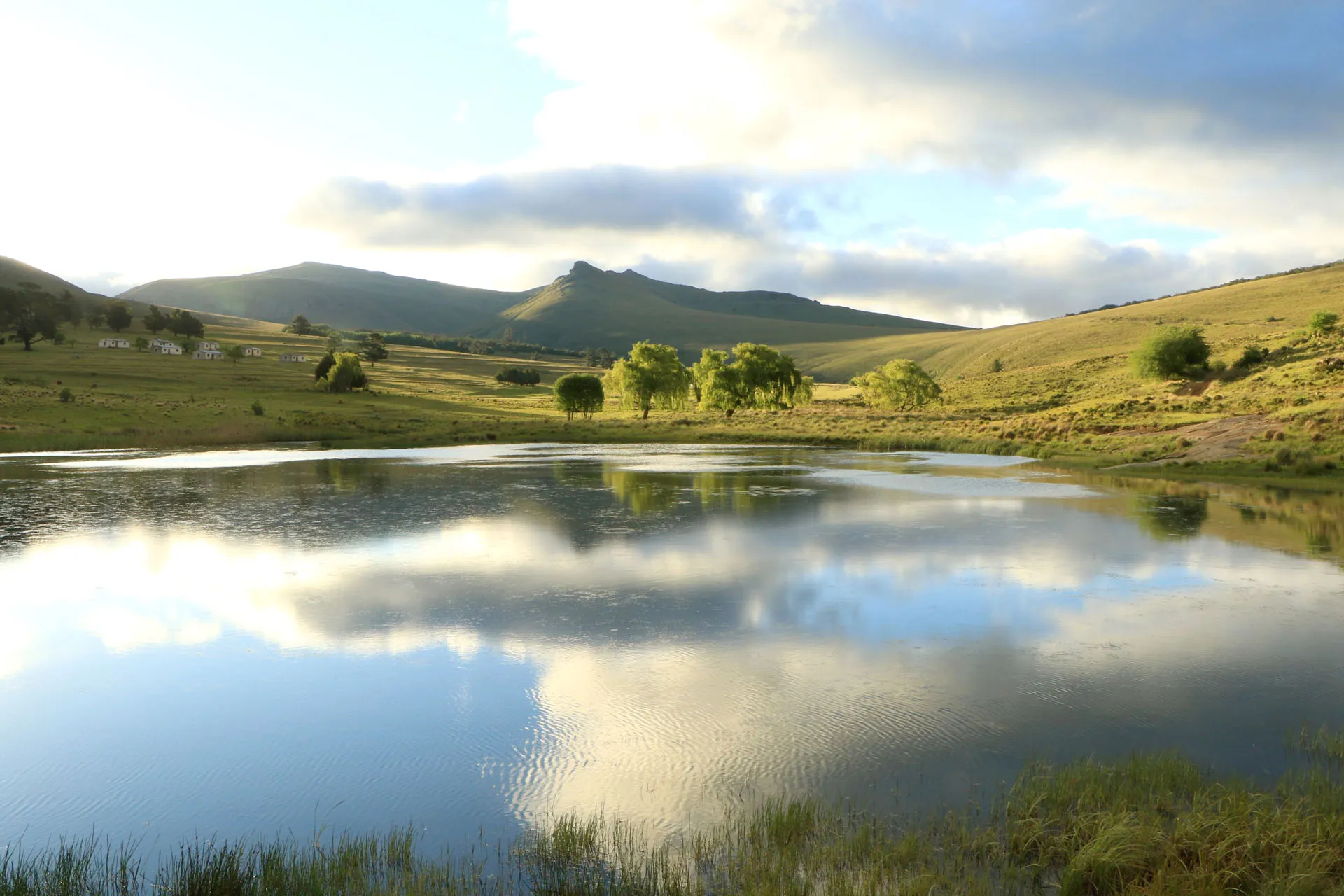 Serene lake with reflections green hills and scattered trees under a partly cloudy sky
