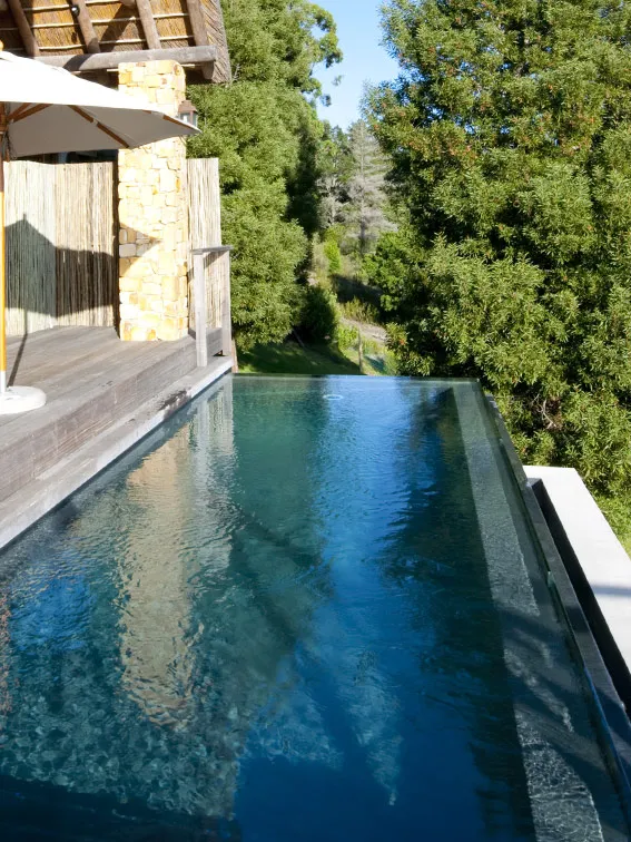 Infinity pool overlooking a wooded area with a wooden deck and umbrella