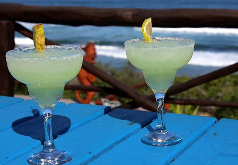 Two margaritas on a blue table with ocean and railing in the background