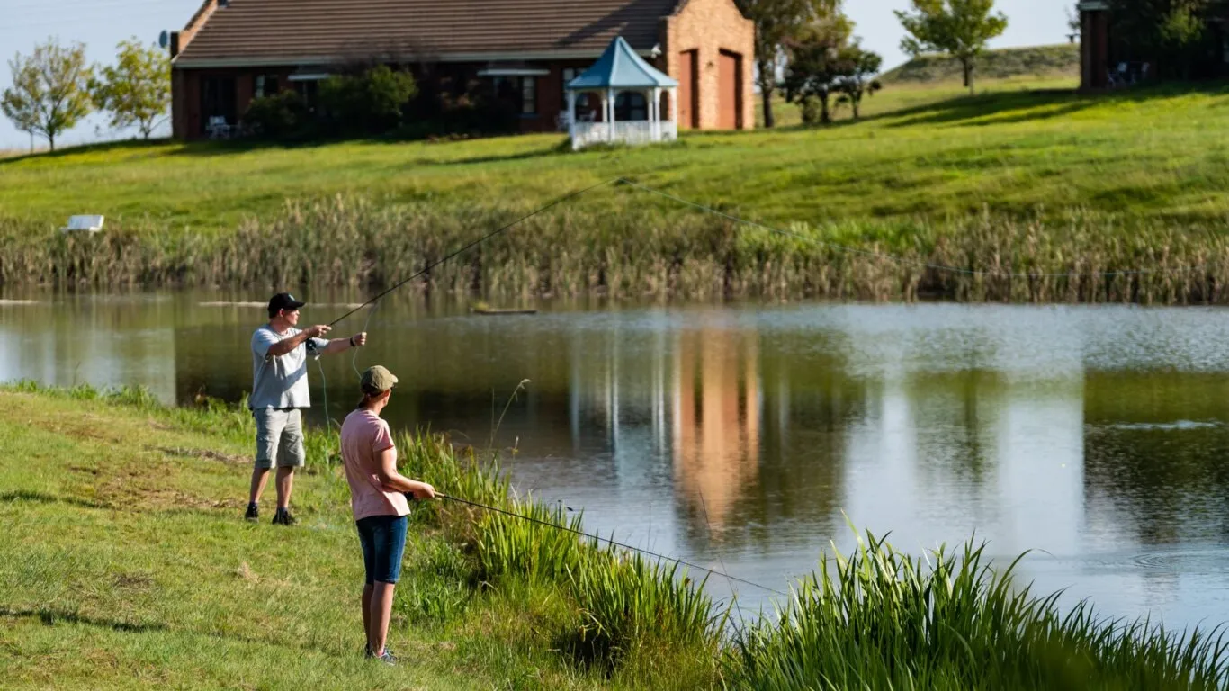 Two people fishing by a pond with a house in the background