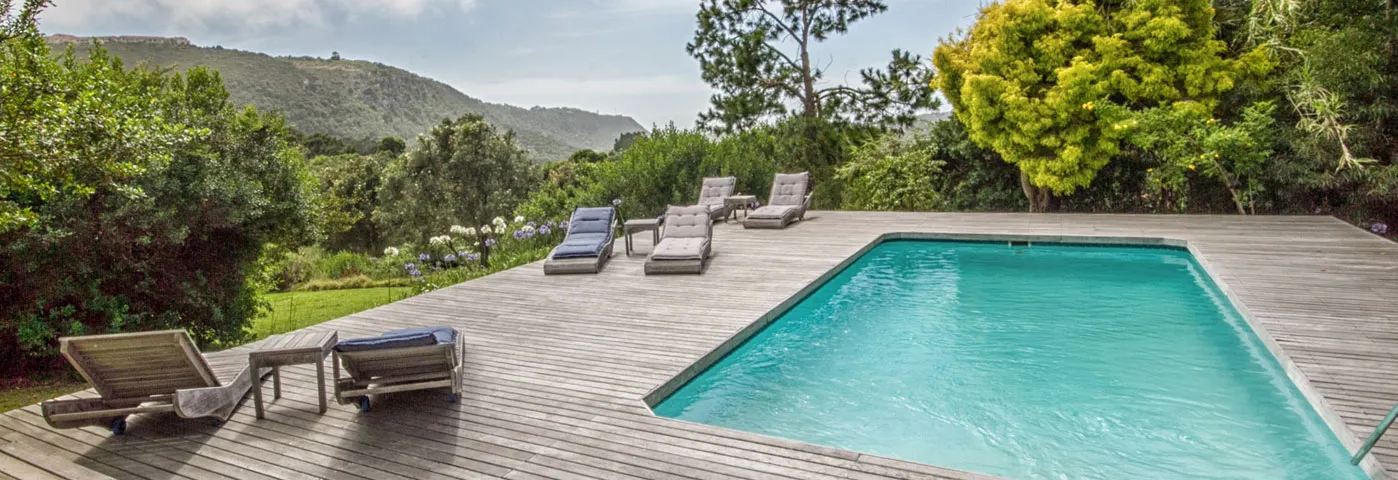 Wooden deck with lounge chairs beside a clear blue swimming pool and greenery