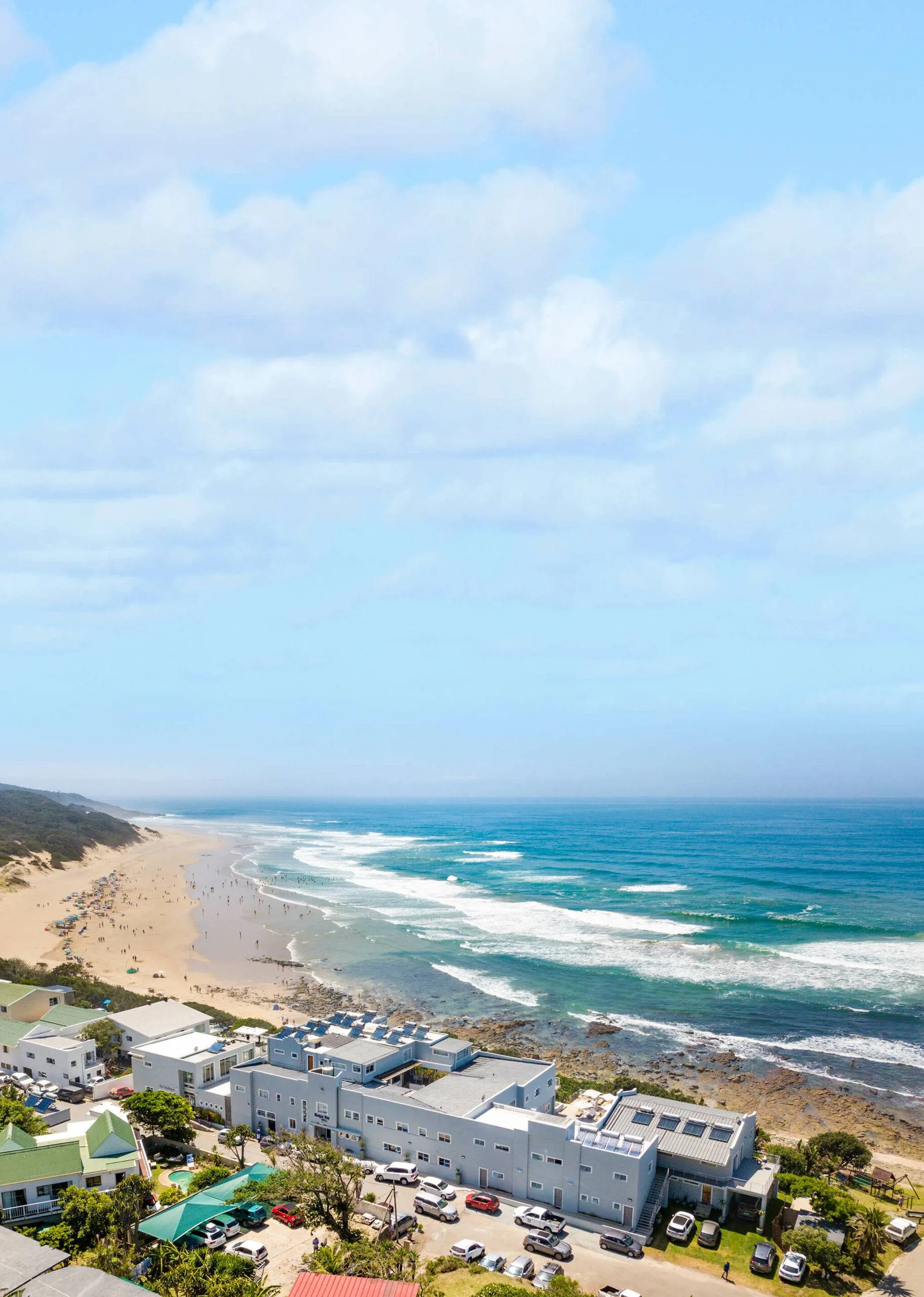 Beachfront buildings with cars parked ocean waves and sandy shore under blue sky