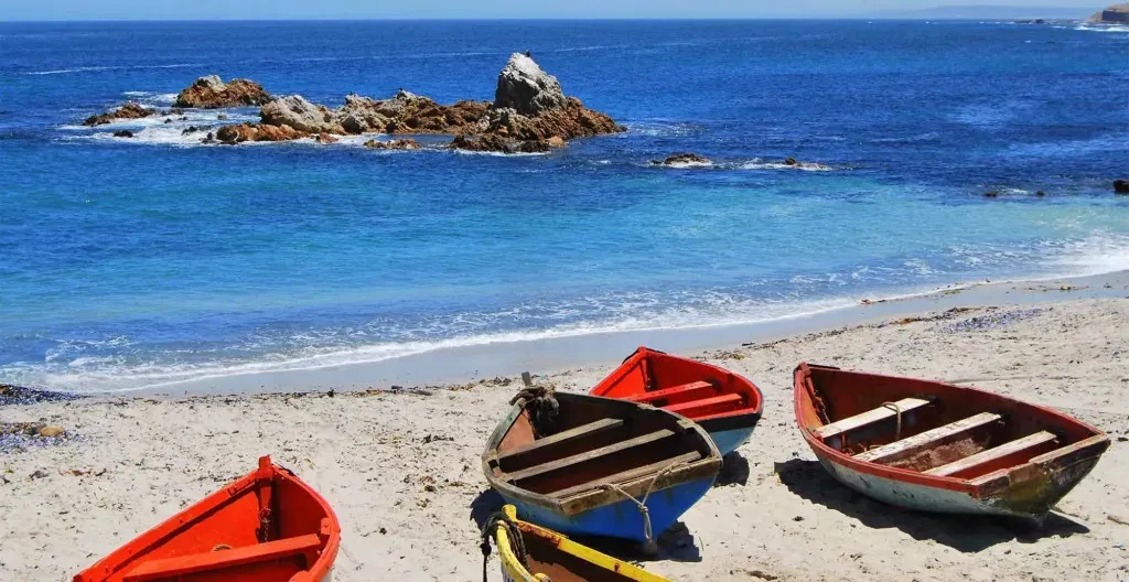 Four colorful boats on a sandy beach with ocean and rocks in the background