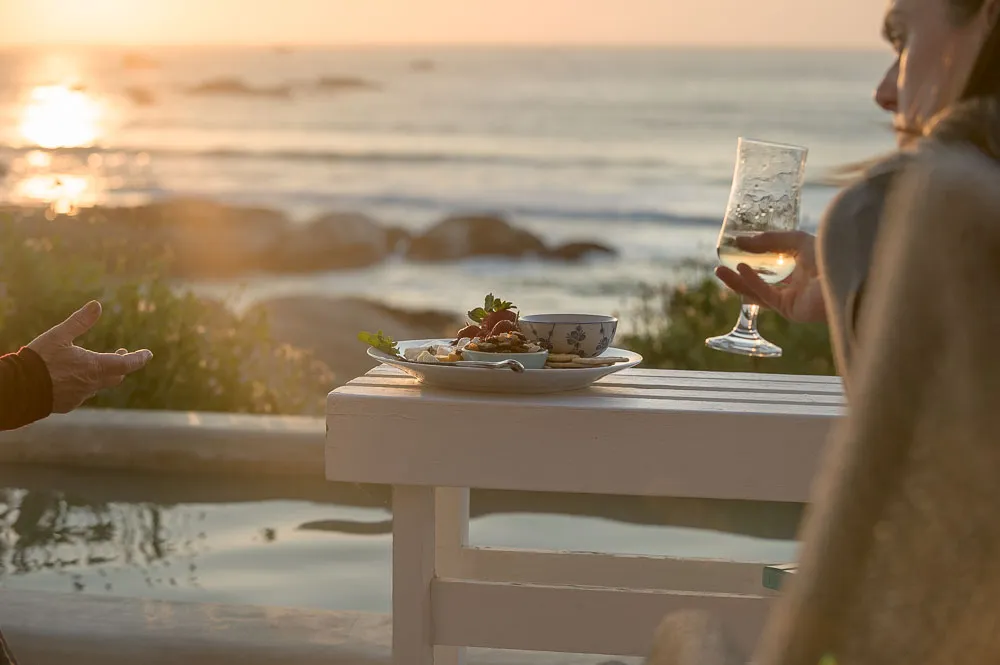 Two people dining outdoors by the ocean at sunset with food and wine