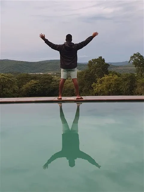 Person standing by pool with arms raised reflecting in water with hills in background