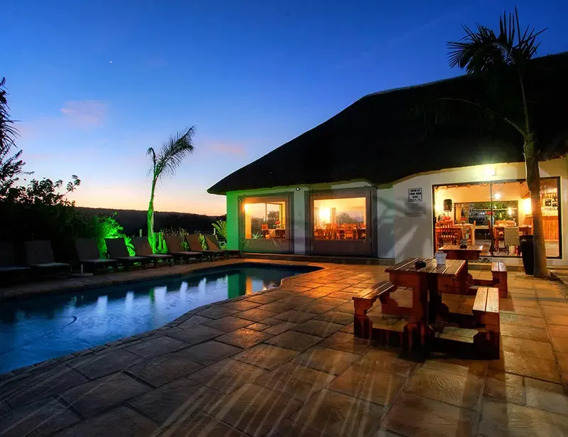 Evening view of a poolside area with wooden benches and a thatchedroof building