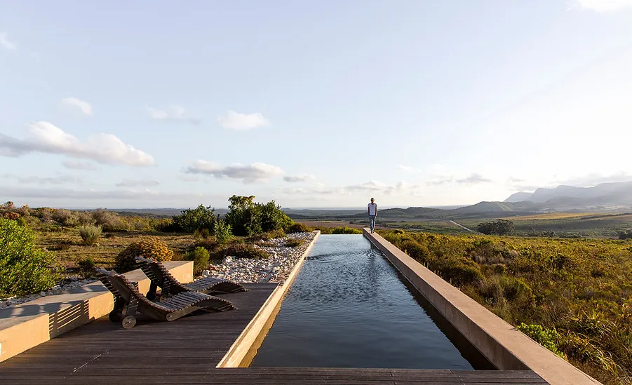 Person standing by a long pool with lounge chairs overlooking a scenic landscape