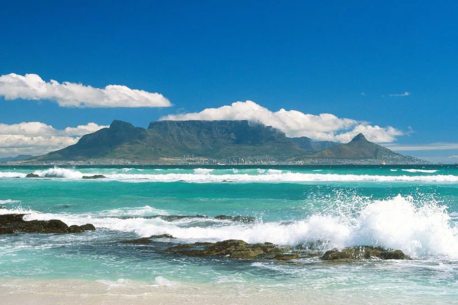 Ocean waves crashing on rocks with a mountain in the background under a blue sky