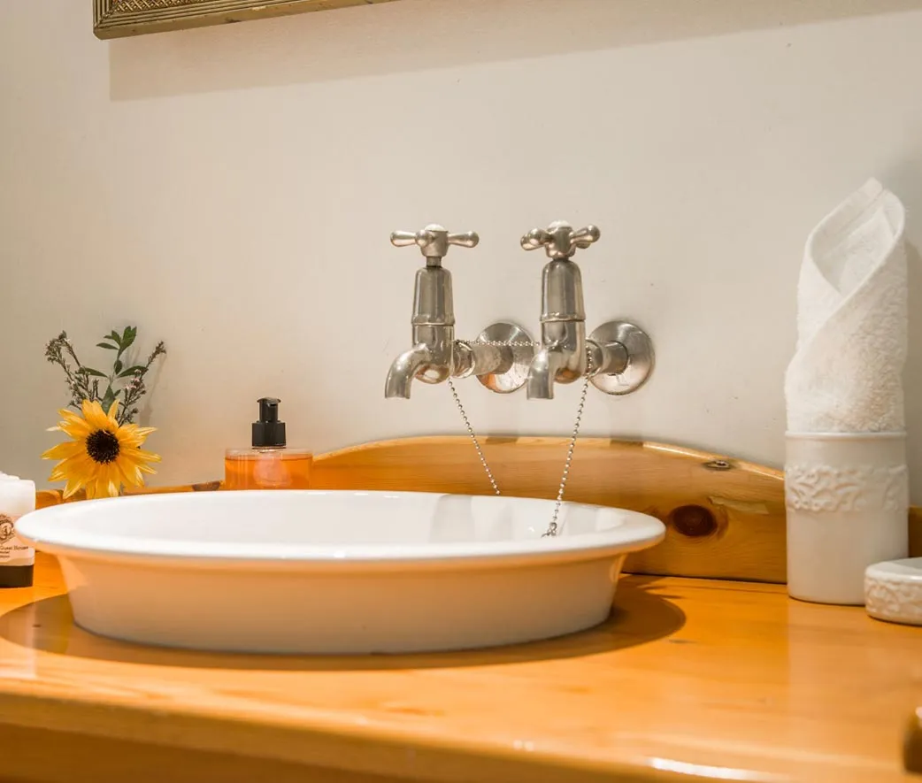 White sink with two metal faucets on a wooden vanity with a flower