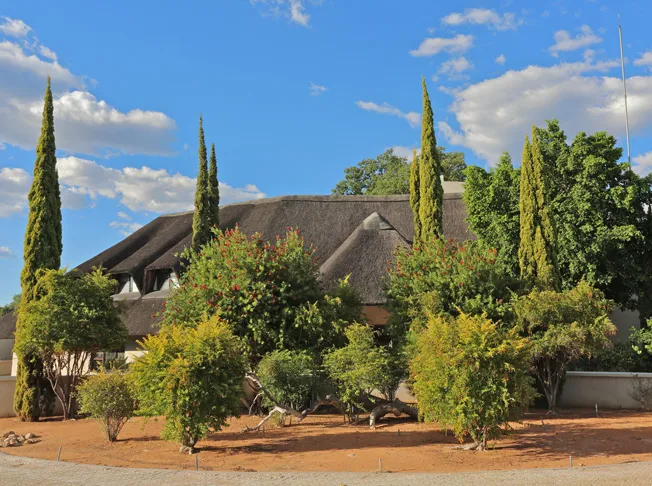 Thatchedroof house surrounded by trees and shrubs under a blue sky with clouds