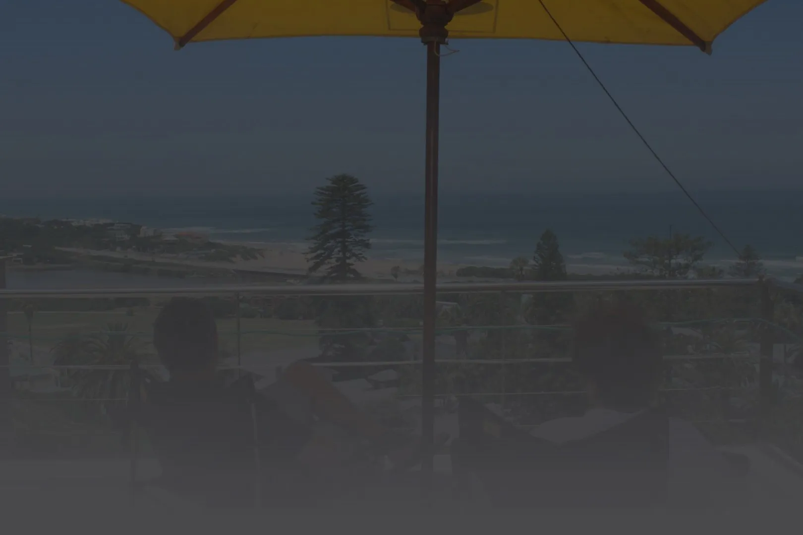 Two people sitting under an umbrella overlooking the ocean from a balcony