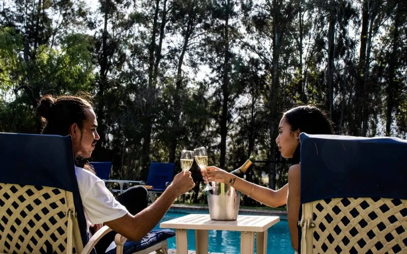 Two people toasting with wine glasses by a pool with trees in the background