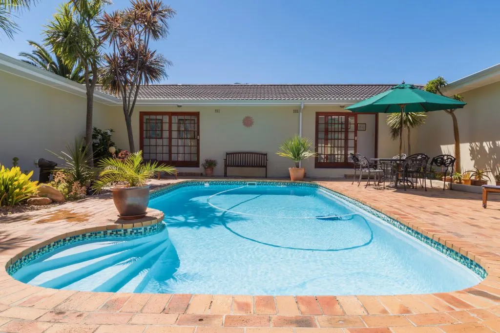 Swimming pool with steps surrounded by plants and patio furniture under a blue sky