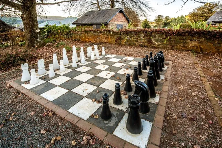 Giant chess set on a large board outdoors with a stone wall background