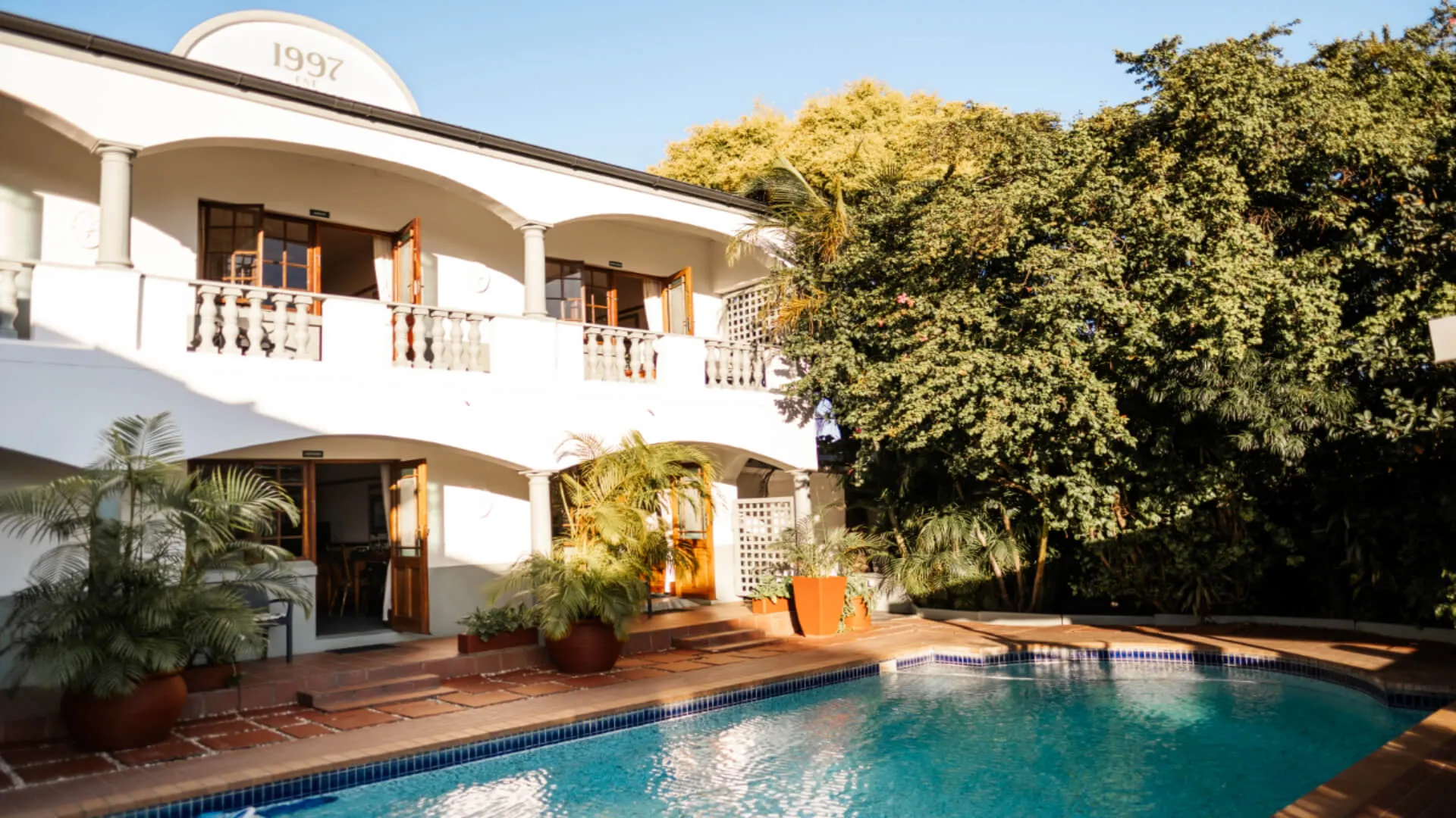 White building with balconies and a swimming pool in front surrounded by greenery