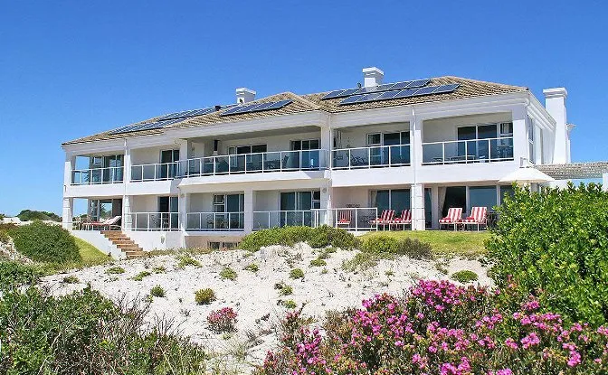 Large white beach house with balconies surrounded by sand dunes and flowers