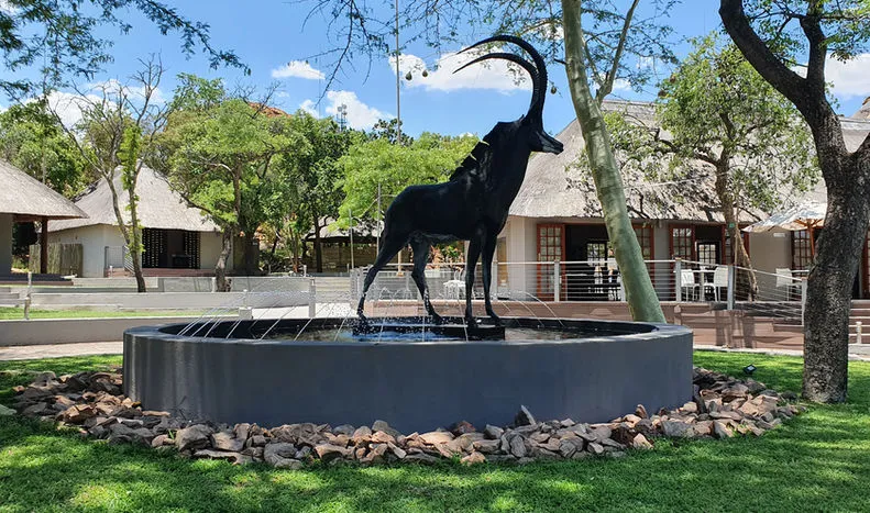 Black antelope statue on a fountain in a grassy park with huts in background