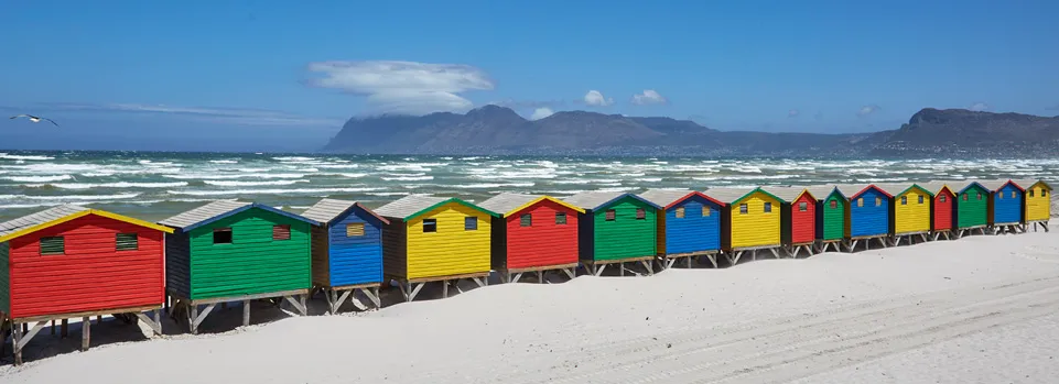 Colorful beach huts on a sandy shore with ocean and mountains in the background