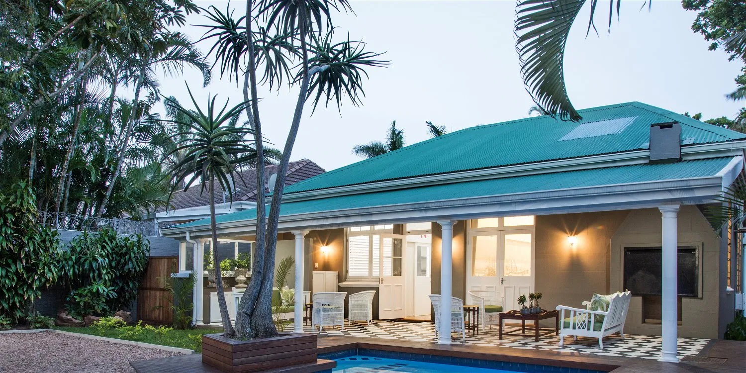 Singlestory house with a green roof white porch and tropical plants around